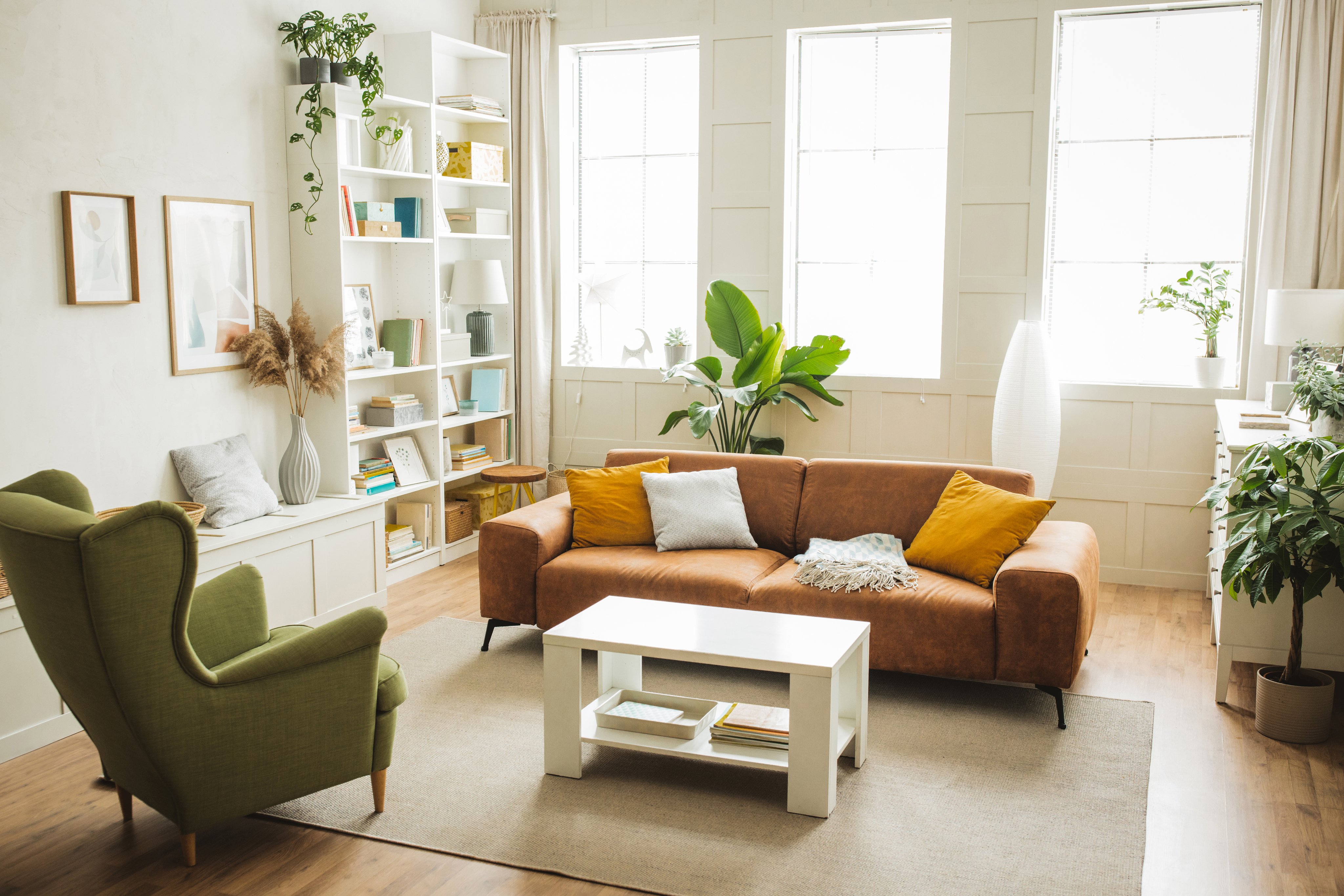 Bright, decluttered living room with a brown leather sofa, mustard and white pillows, a green wingback chair, a white coffee table, white bookshelves, plants, a light wood floor and tall windows with sheer curtains