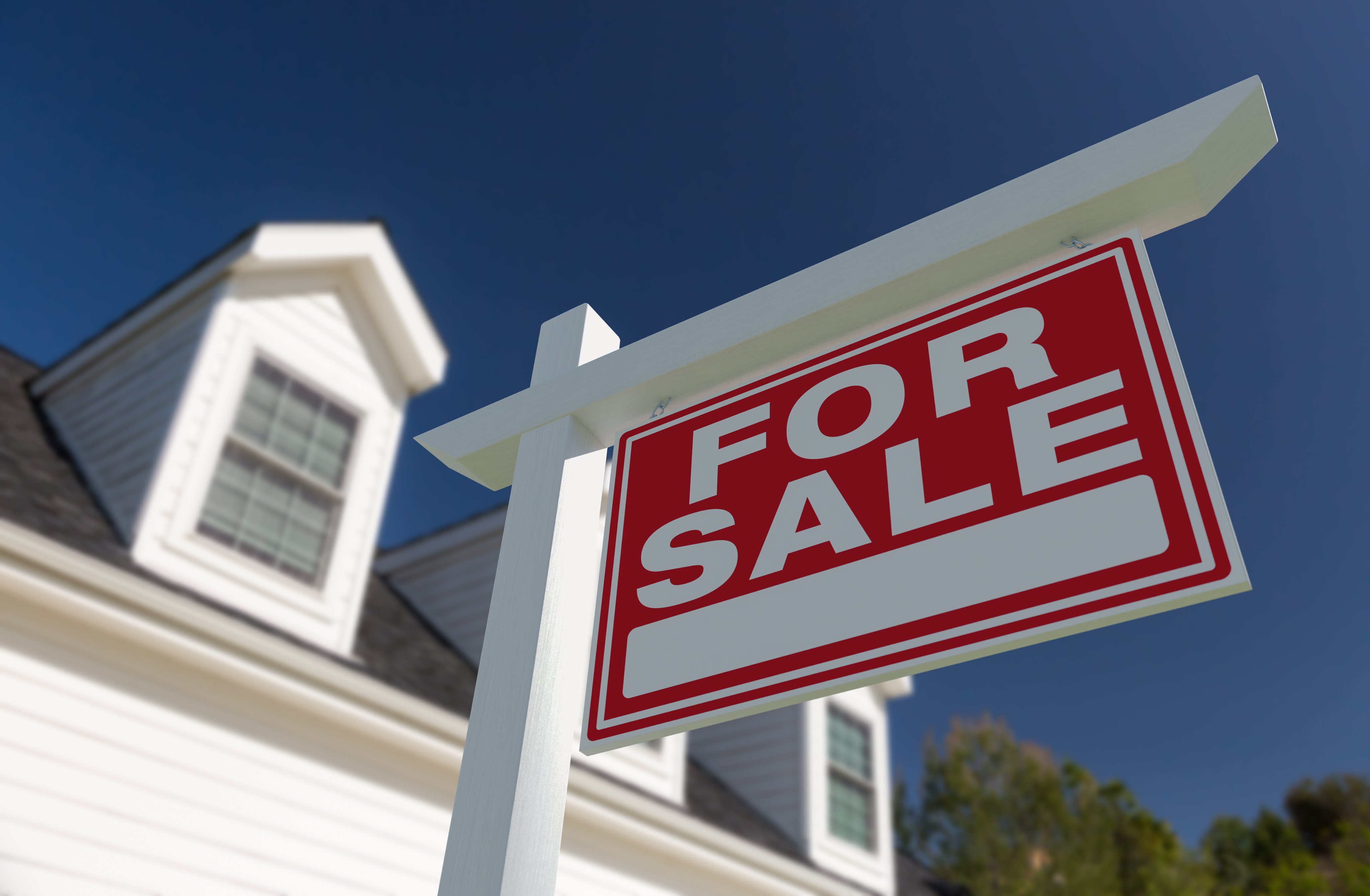 A red and white FOR SALE real estate sign in front of a house against a blue sky.