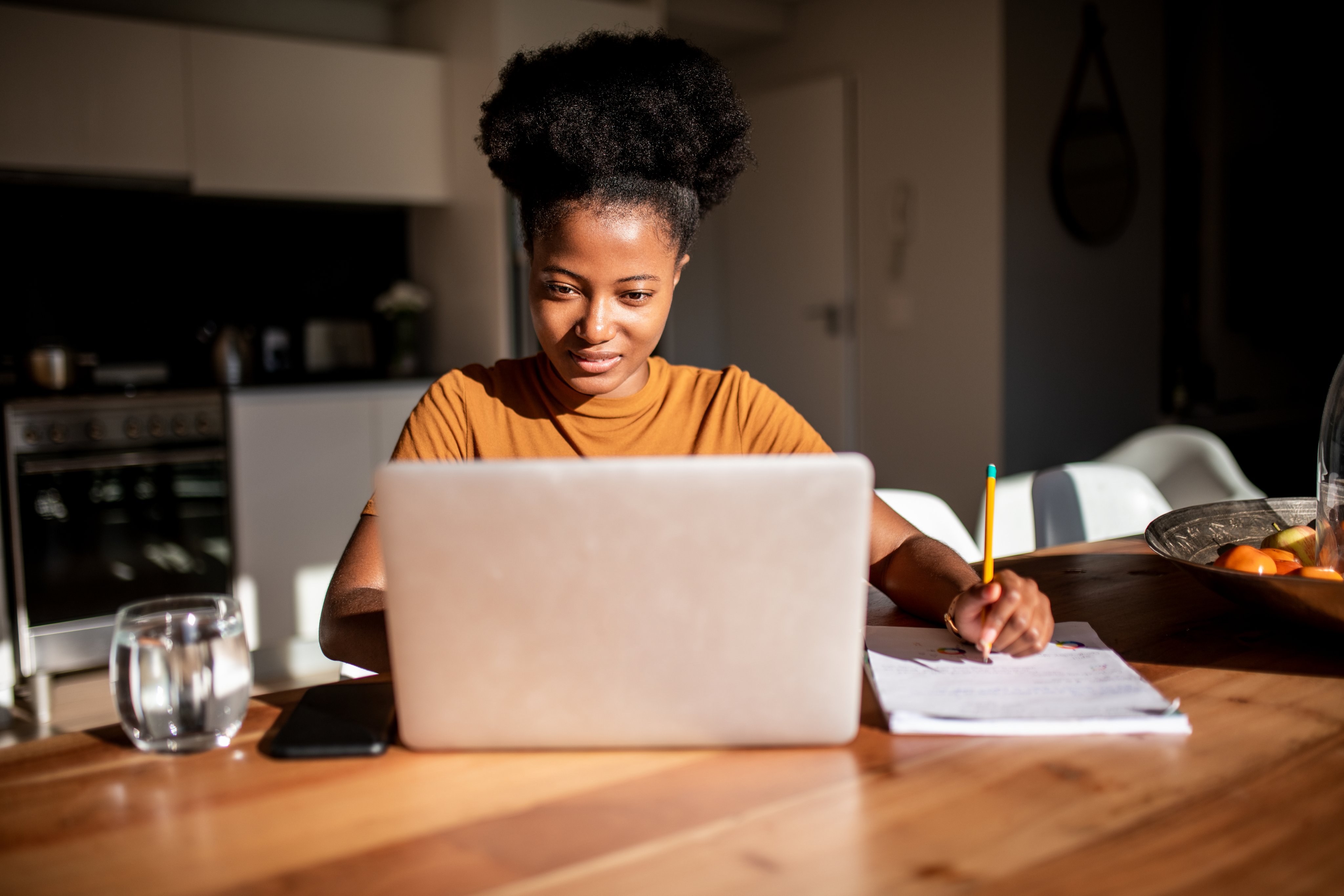 Focused learner studying on a laptop with notes at a kitchen table in natural light