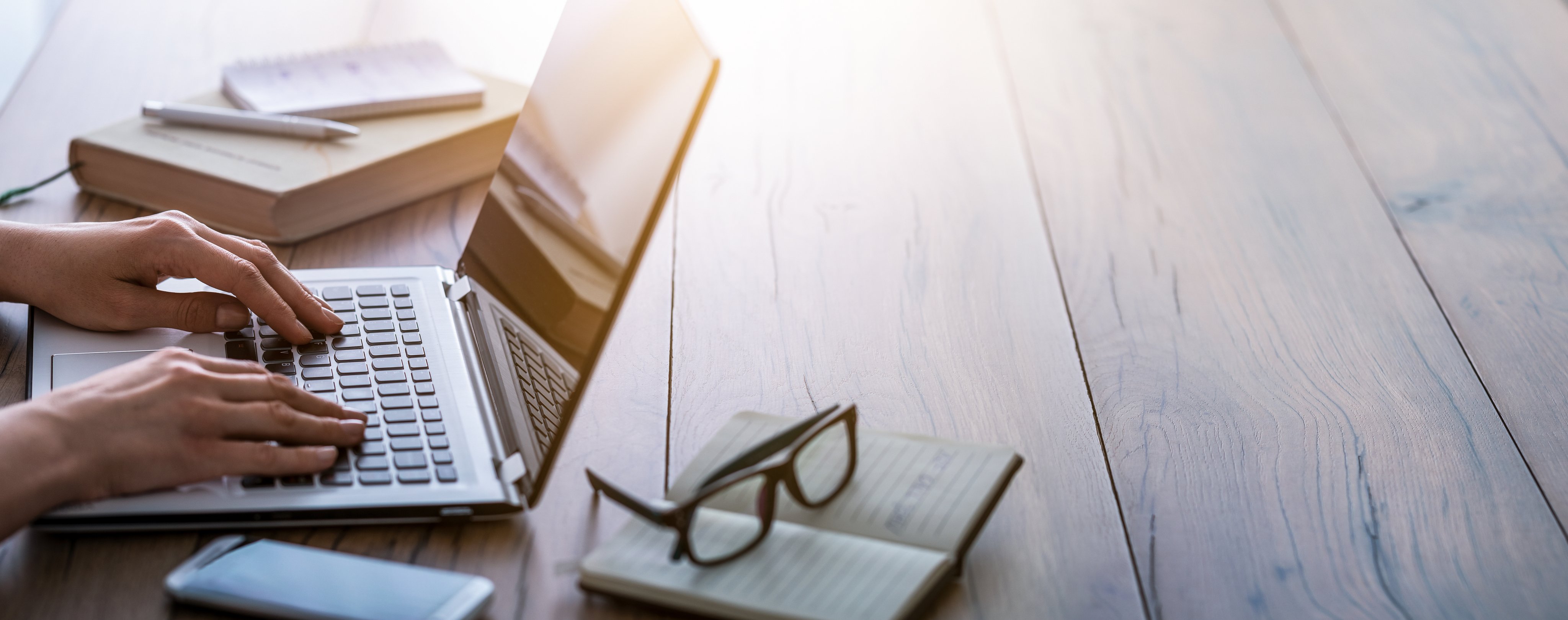 Study desk with a laptop, stacked books, notepad, and glasses in warm natural light