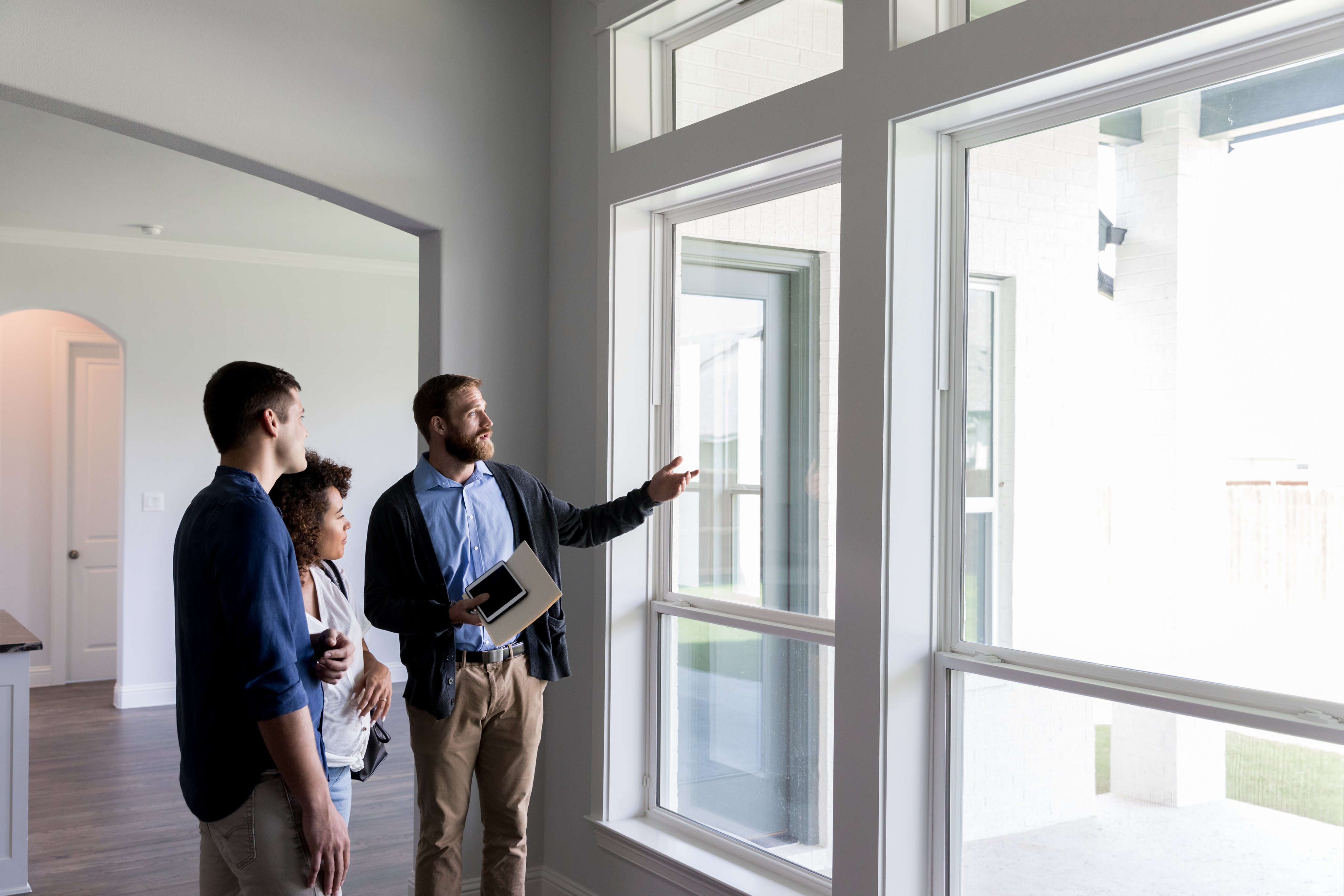 Real estate agent holding a tablet and pointing out large windows to a young couple during a home showing