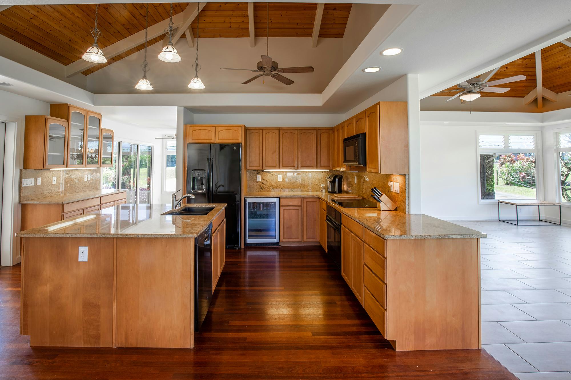 Kitchen with wood cabinets, granite counters, an island with a sink, a black refrigerator, pendant lights, a wood-beamed vaulted ceiling, dark hardwood floors and windows along the right wall