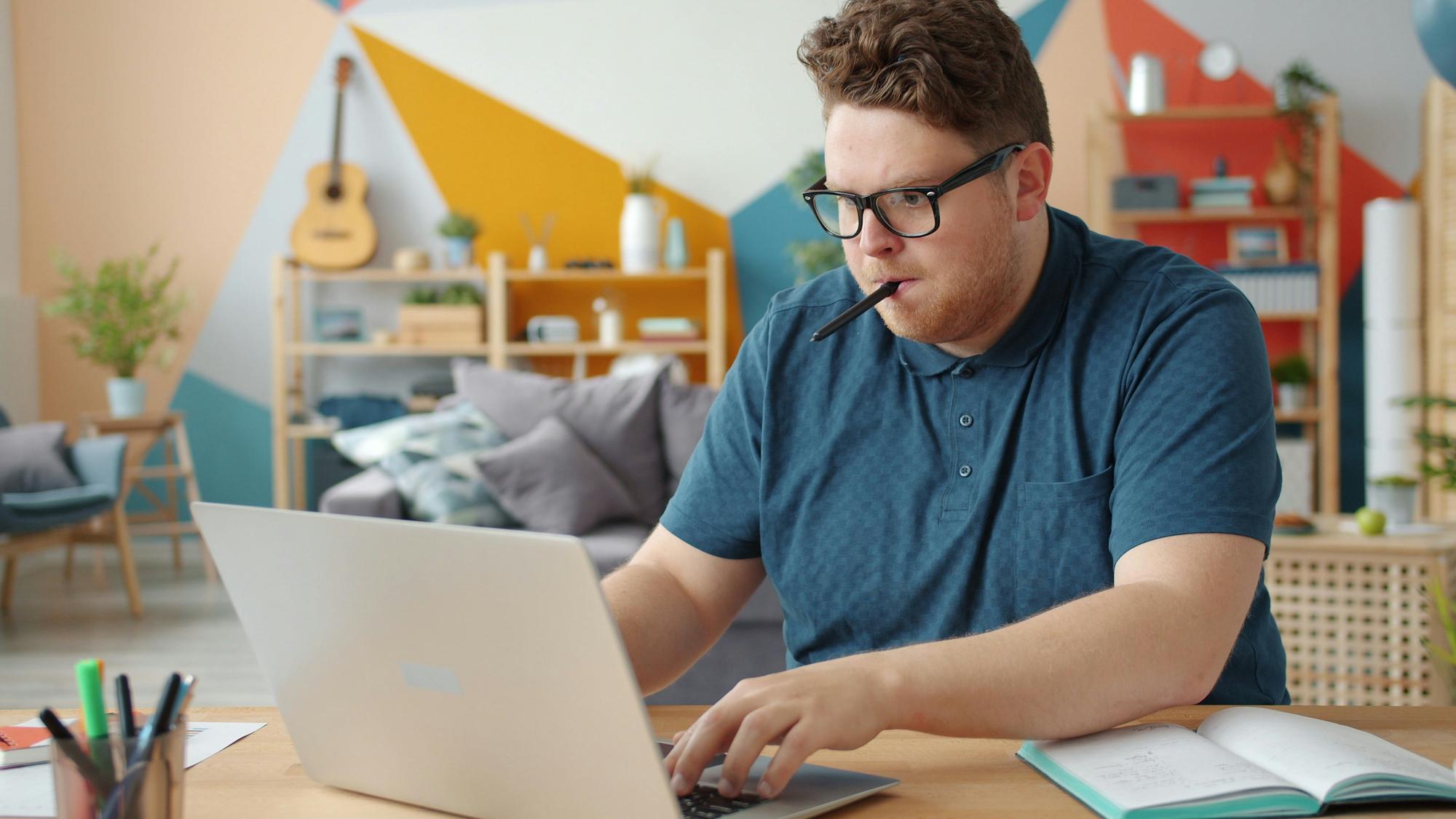 Adult learner studying at a laptop at home with an open notebook of handwritten notes beside him.